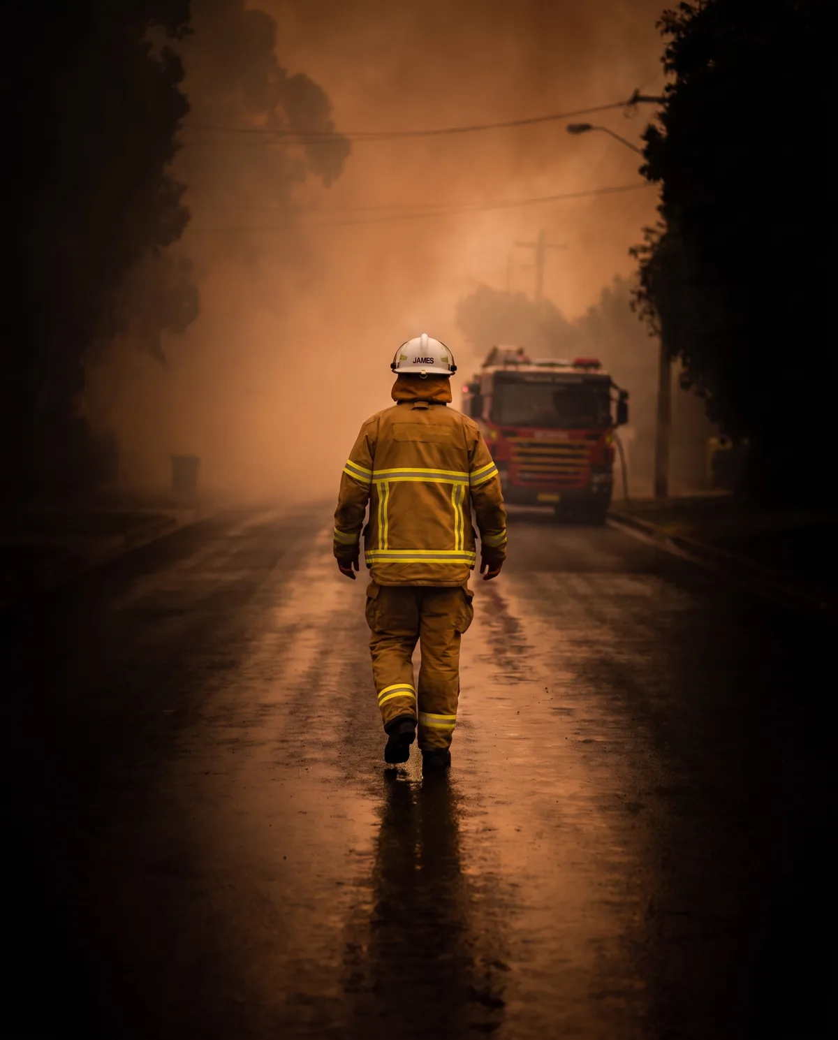 A firefighter in Australian turnout gear walking toward a fire appliance in smoky dusk light